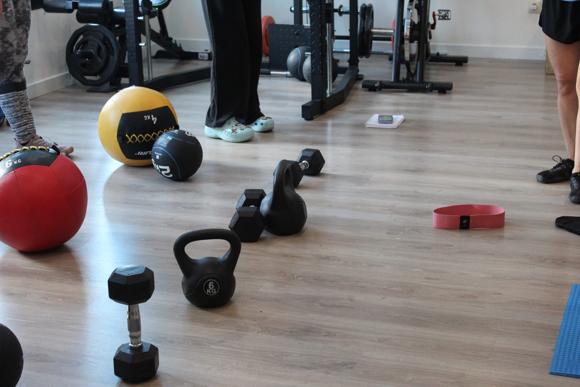 Clase de entrenamiento funcional: pesas rusas, mancuernas y balones medicinales en el suelo de la sala, con las alumnas al fondo